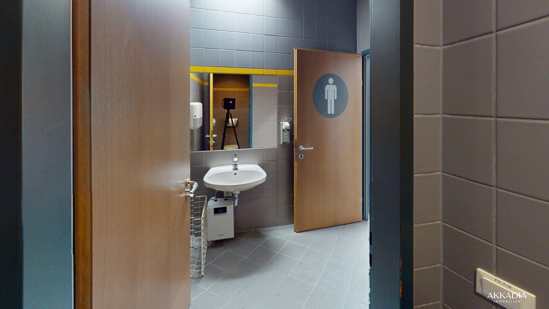 Öffentliche Toilette mit grauen Fliesen, Waschbecken und einer Tür mit Männchen-Symbol.