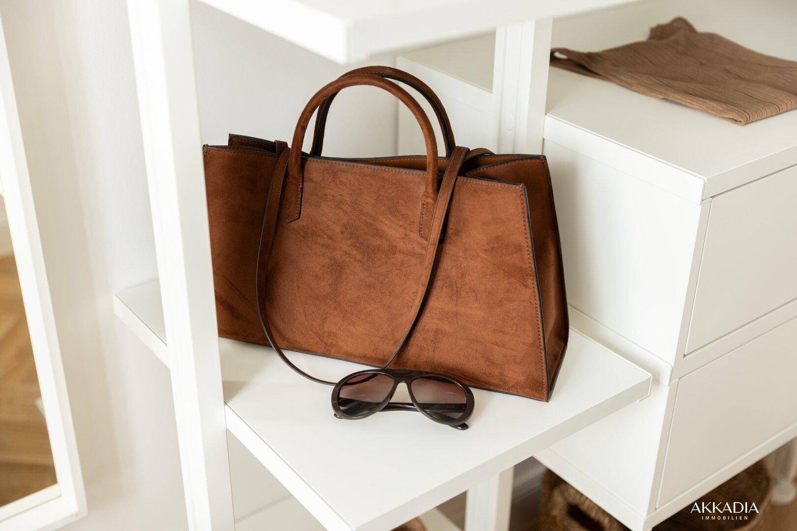 Nahaufnahme einer braunen Handtasche und Sonnenbrille auf einem Regal im Schlafzimmer.