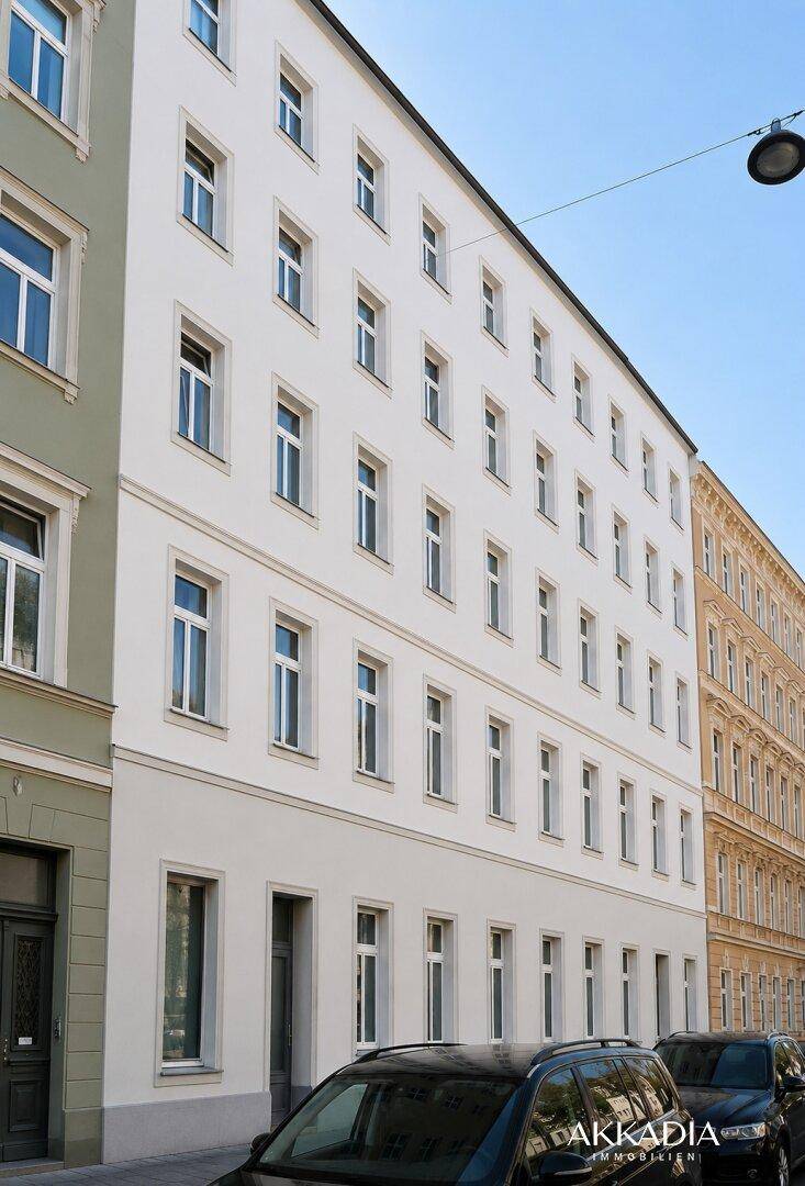 Ansicht einer weißen Wohnhausfassade mit zahlreichen Fenstern und parkenden Autos auf der Straße.