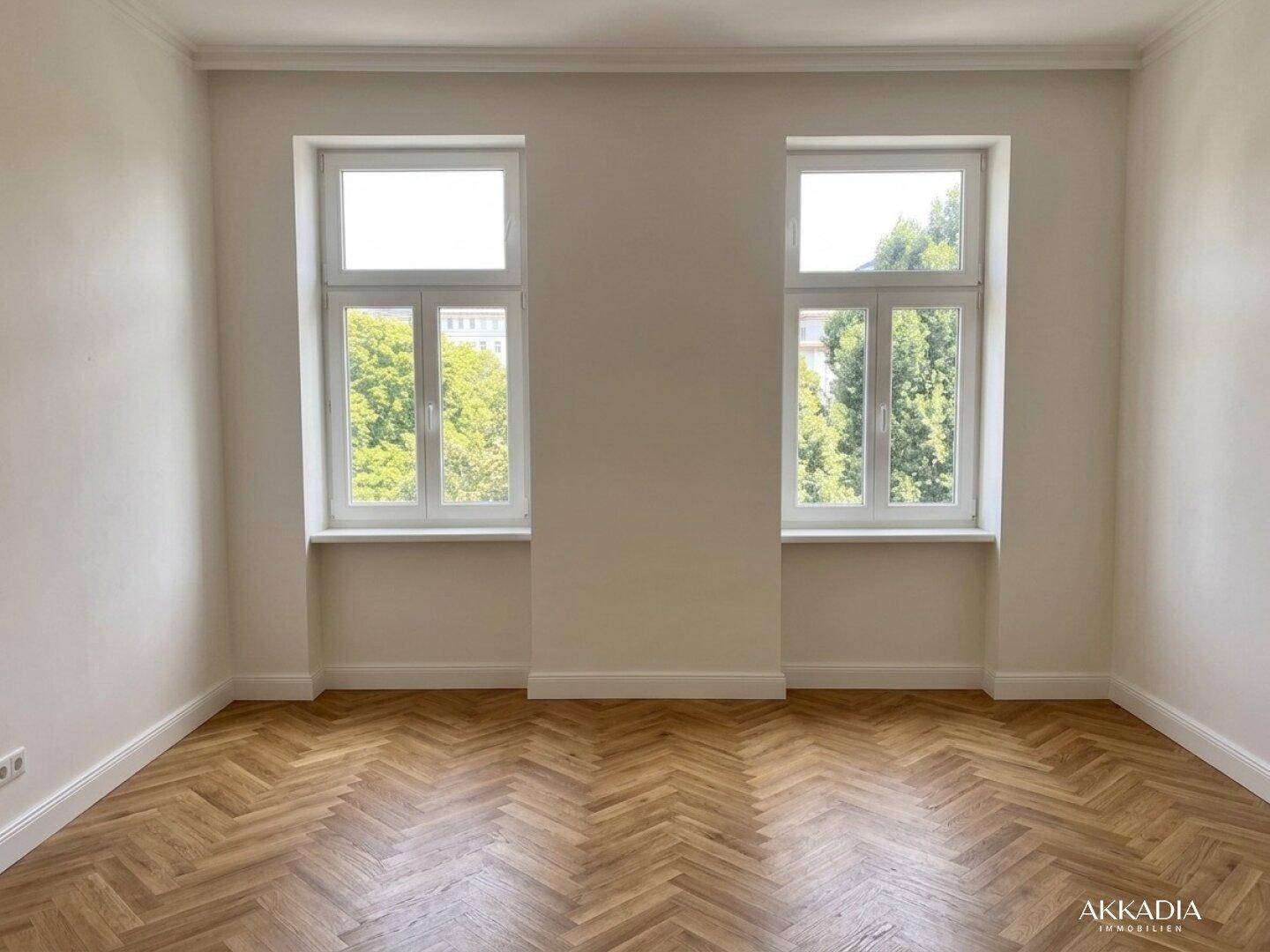 Heller, leerer Raum mit Fischgrätparkett und zwei großen Fenstern, die viel Licht hereinlassen.