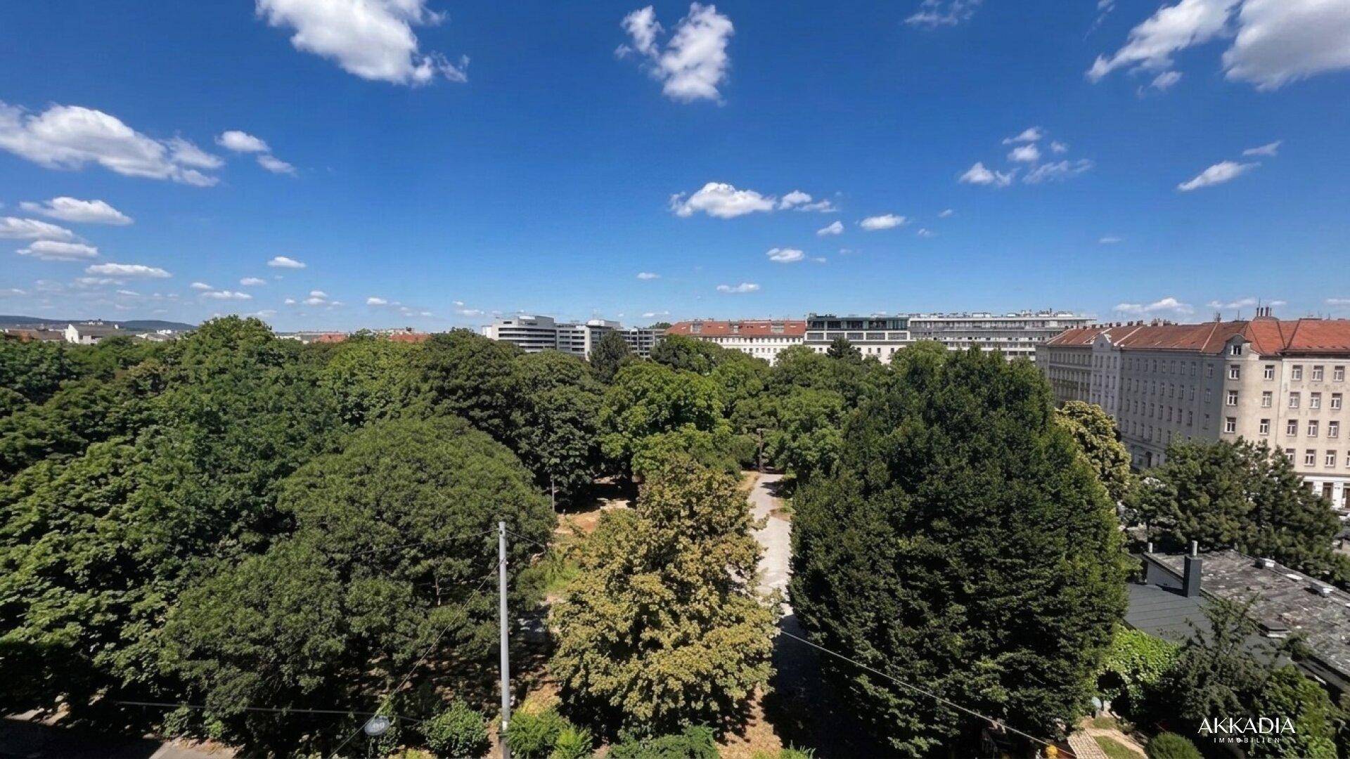Weitläufiger Blick über eine grüne Parklandschaft und die Dächer der Stadt unter blauem Himmel.