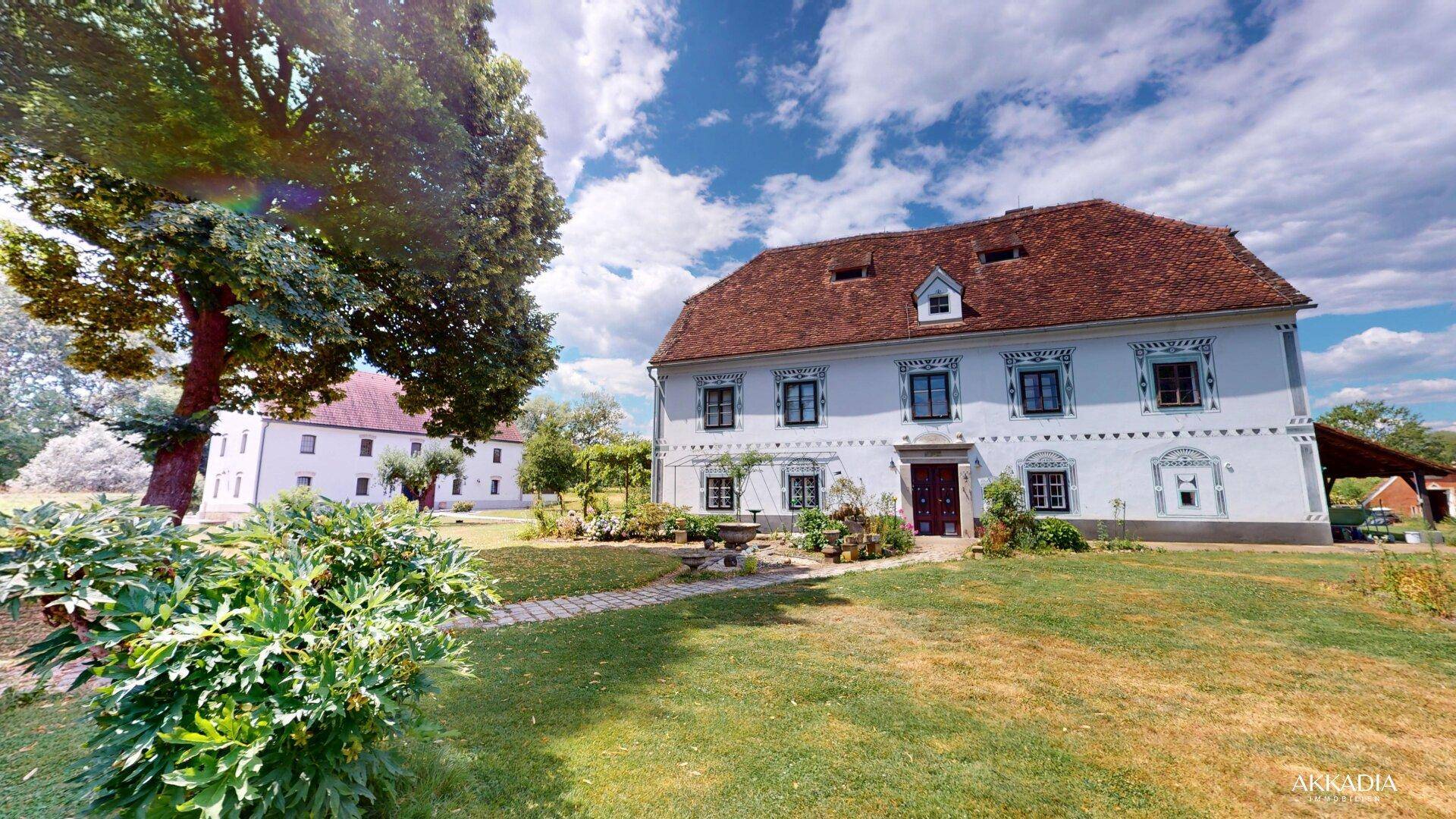 Historisches Gebäude mit weißer Fassade, umgeben von einem weitläufigen Garten mit alten Bäumen.
