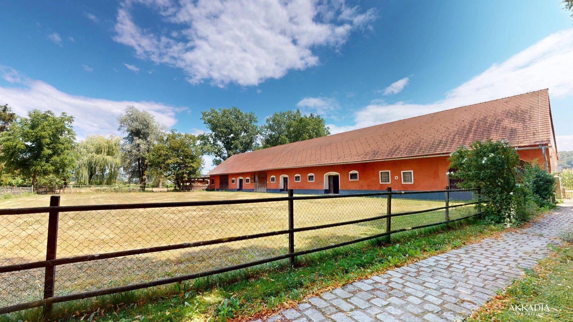 Langes Stallgebäude mit rotem Dach und Zaun, umgeben von weitläufigen Grünflächen.
