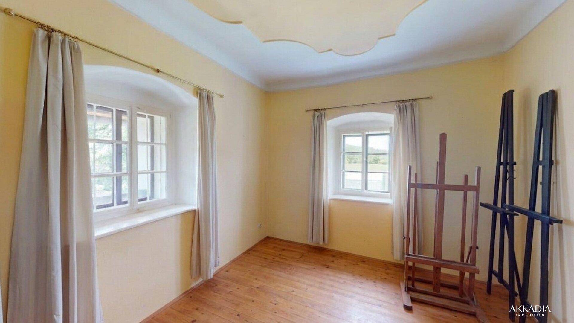 Helles Zimmer mit Holzboden, zwei Fenstern und einer Staffelei, ideal als Schlaf- oder Arbeitszimmer.