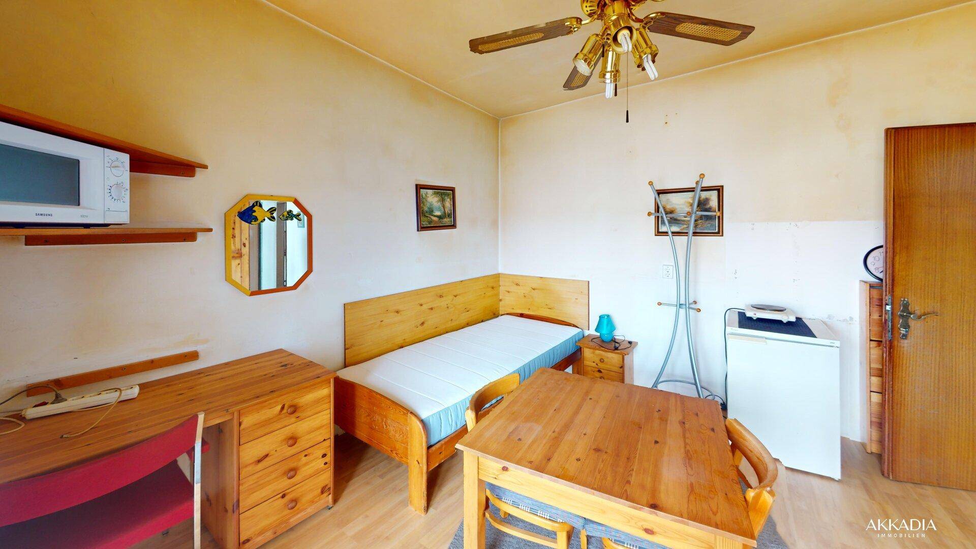 Schlafzimmer mit Einzelbett, Holzmöbeln, Mikrowelle und einem Deckenventilator.