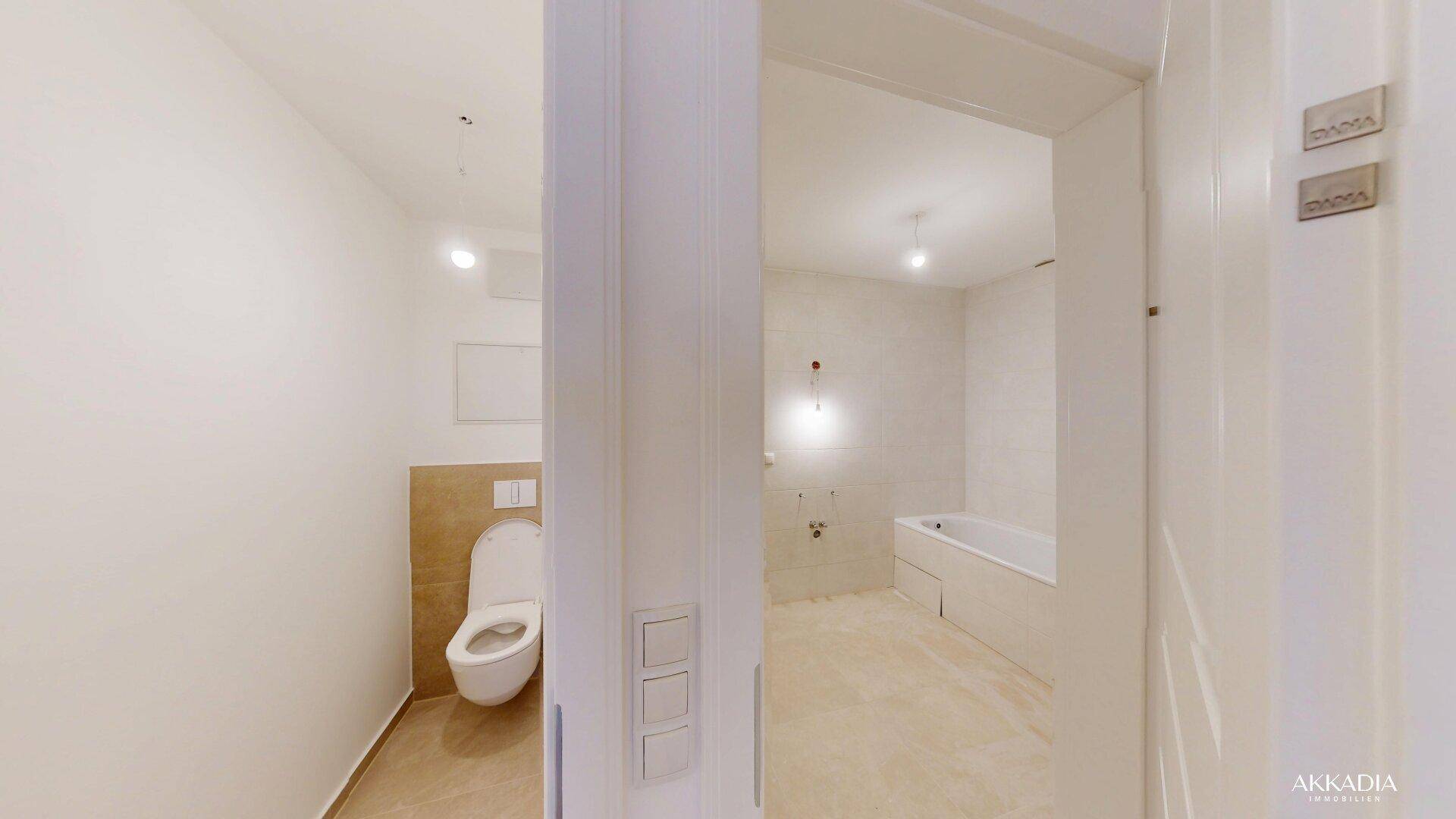 Blick vom Flur in ein separates WC mit Wand-WC und ein Badezimmer mit Badewanne.