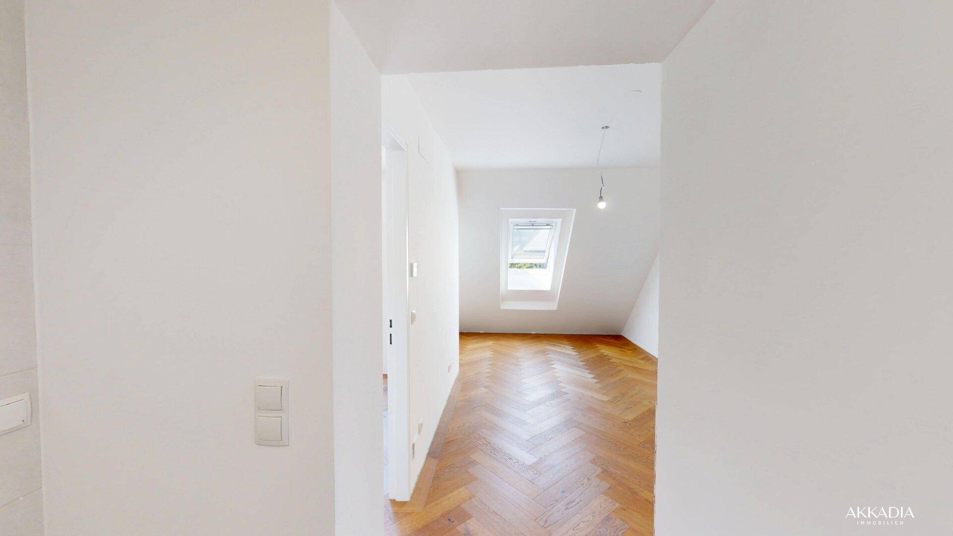 Heller Flur mit Fischgrätparkett und weißen Wänden, der den Blick in ein Zimmer mit Dachfenster freigibt.