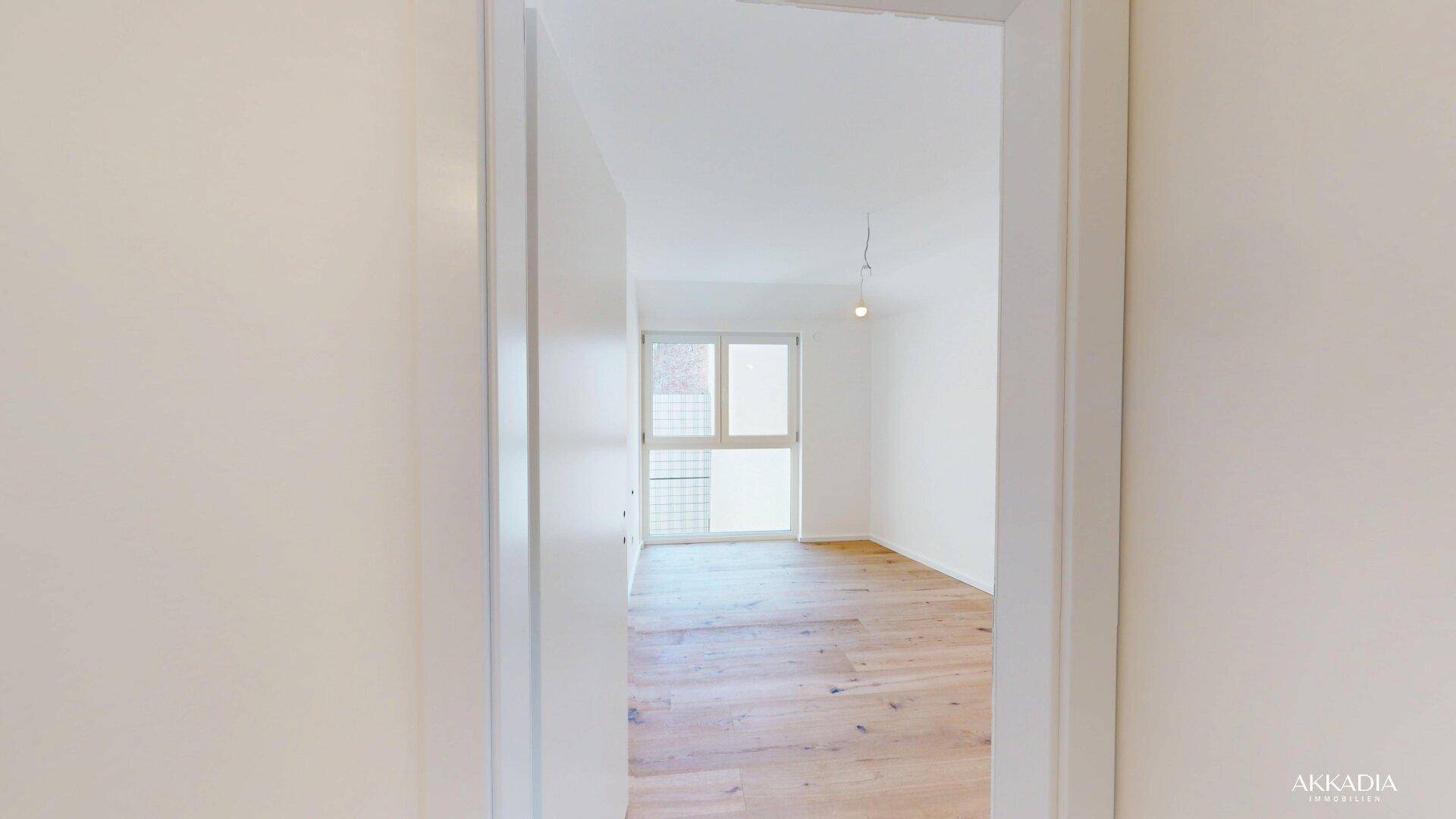 Blick durch eine Tür in ein helles Zimmer mit Holzparkettboden und großem Fenster.