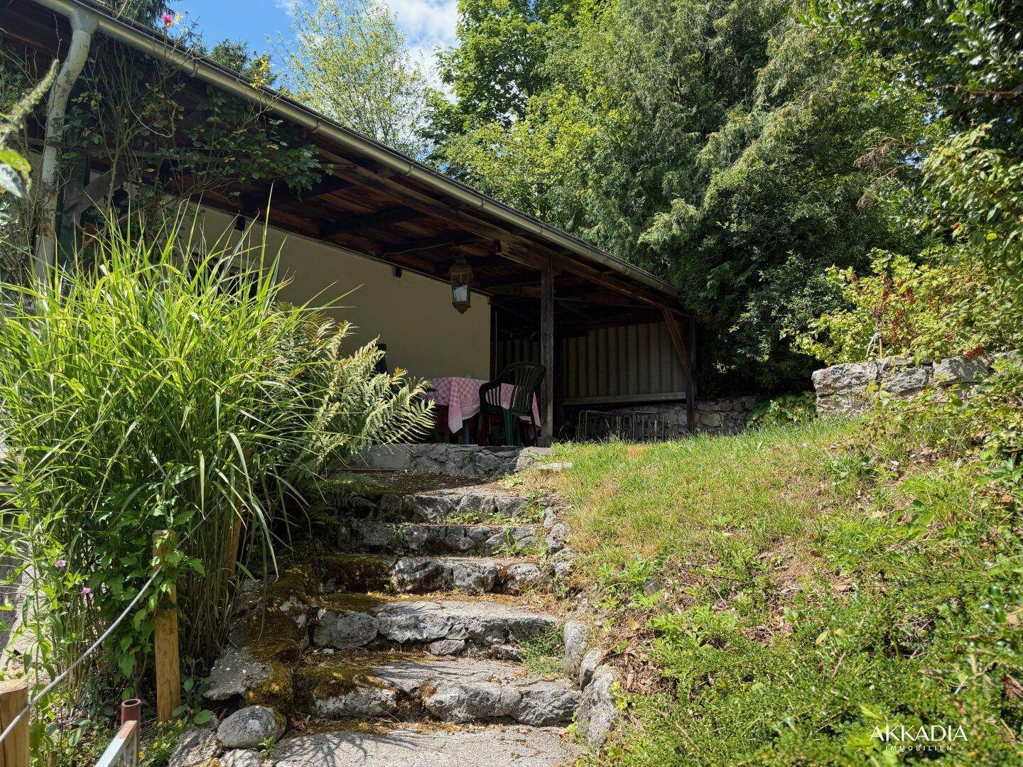 Überdachte Terrasse mit Steintreppe, umgeben von viel Grün und Pflanzen.