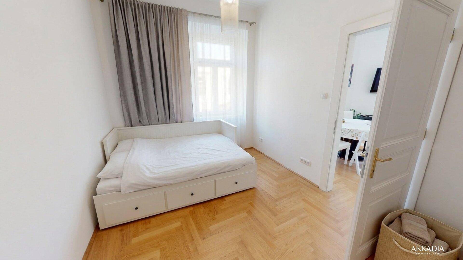 Helles Schlafzimmer mit Einzelbett, Fischgrätparkett und Fenster mit grauen Vorhängen.