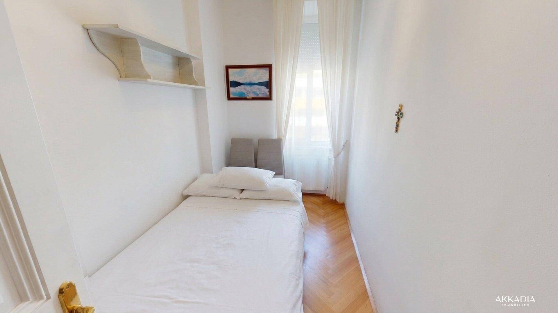 Schlichtes Schlafzimmer mit Einzelbett, weißen Wänden und hellem Fenster.