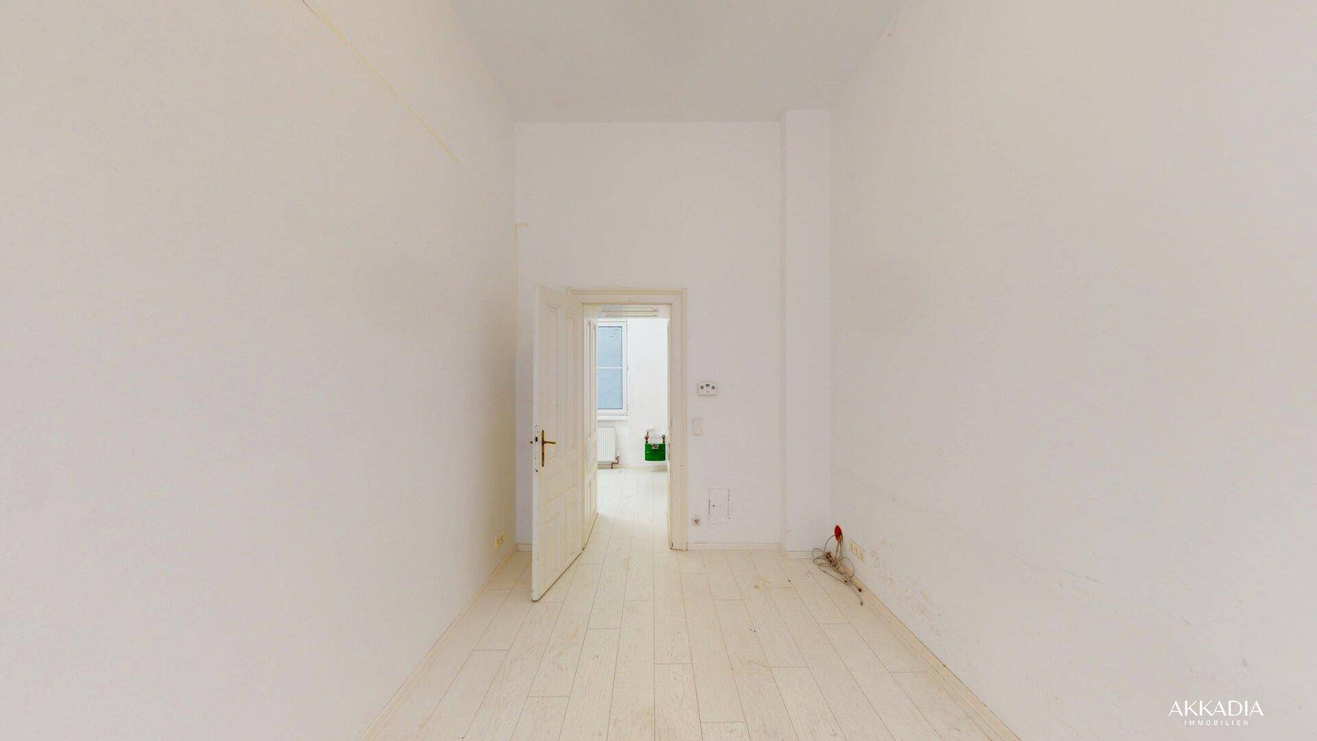 Langer, heller Raum mit weiß gestrichenen Wänden, hellem Dielenboden und einer offenen Tür zu einem weiteren Zimmer.