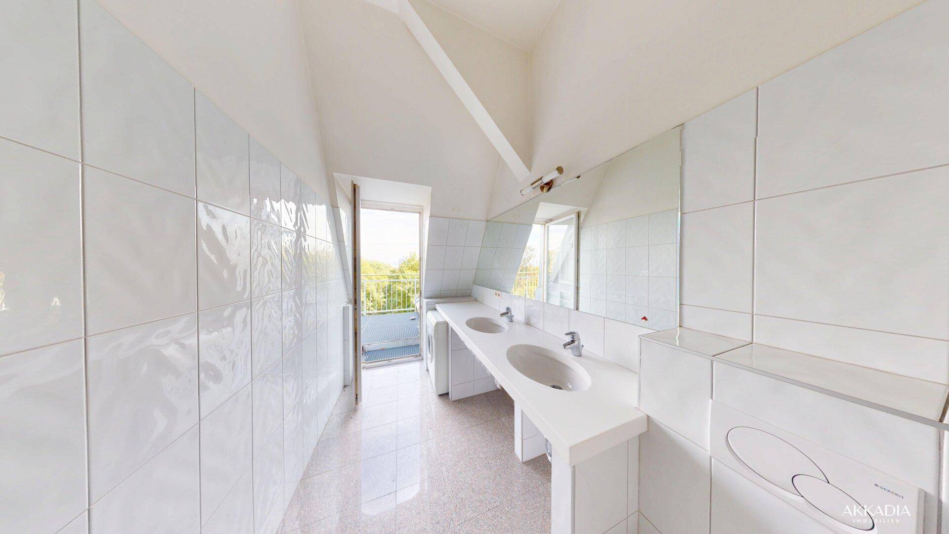 Helles Badezimmer mit weißen Fliesen, Doppelwaschbecken und Zugang zu einem kleinen Balkon.