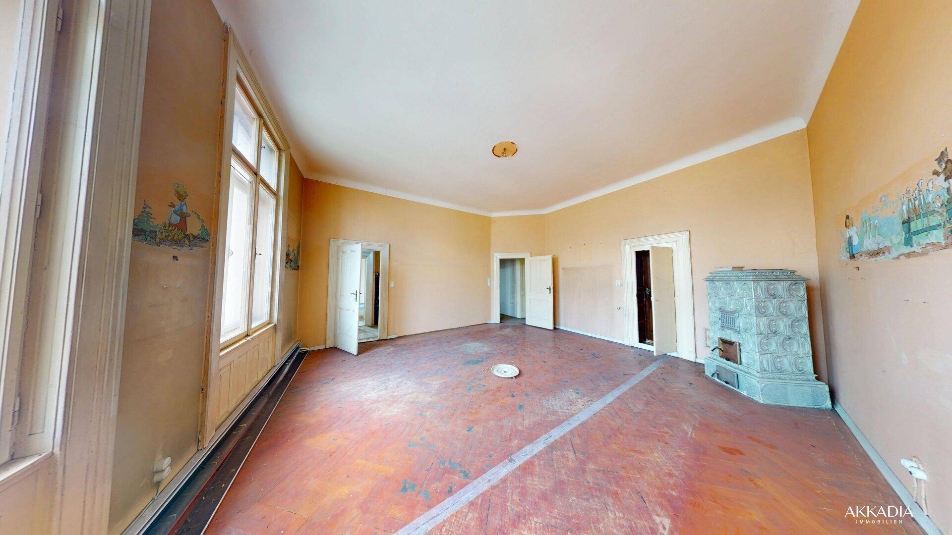 Großes Zimmer mit Kachelofen, Parkettboden und Wandmalereien, das Renovierungsbedarf aufweist.