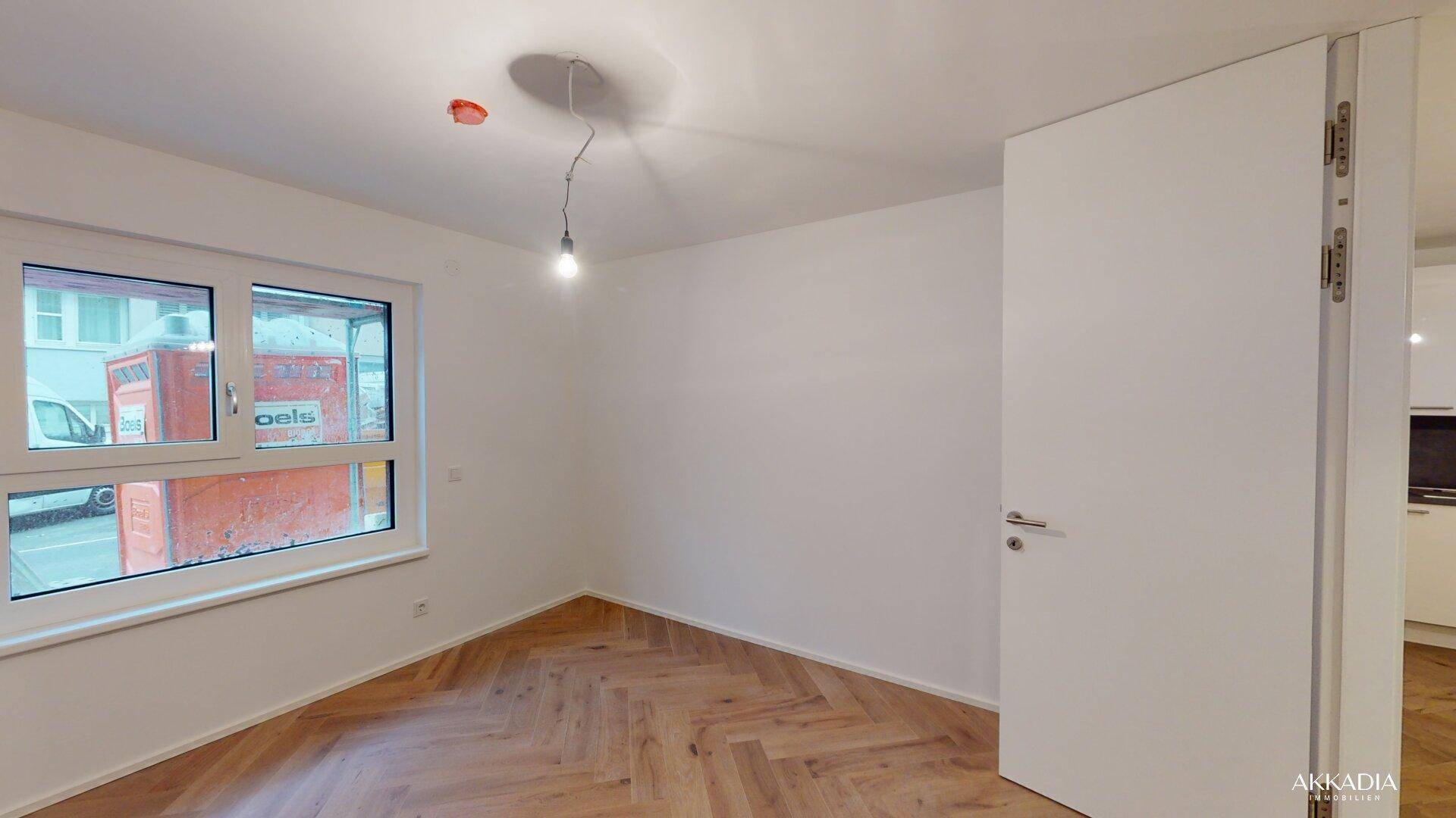 Helles Zimmer mit Fischgrätparkett und weiß gestrichenen Wänden, mit Fenster zur Straße.