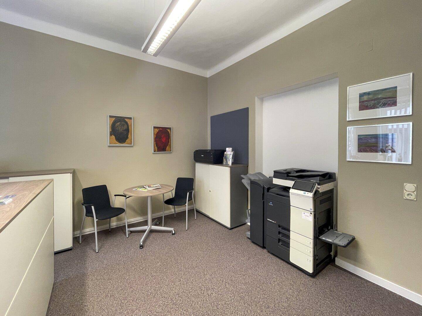 Wartebereich in einem Büro mit zwei Stühlen, kleinem Tisch und einem großen Multifunktionsdrucker.