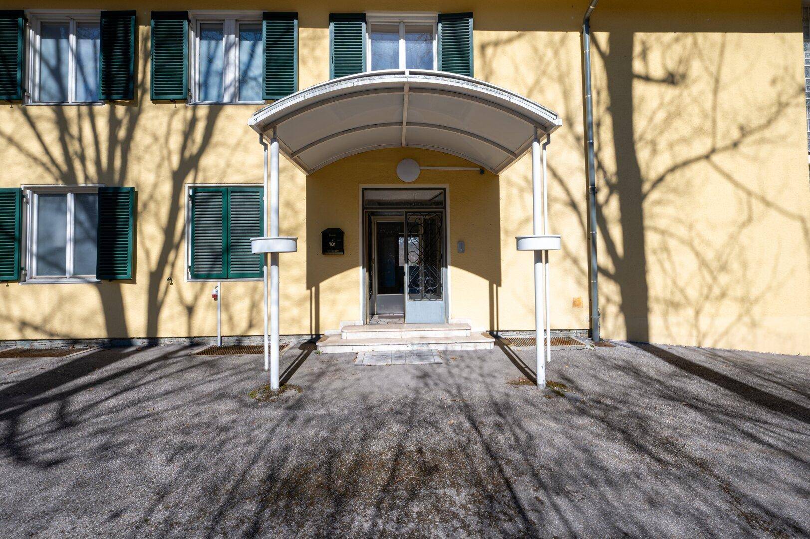 Eingangsbereich eines gelben Gebäudes mit Vordach und grünen Fensterläden an sonnigem Tag.