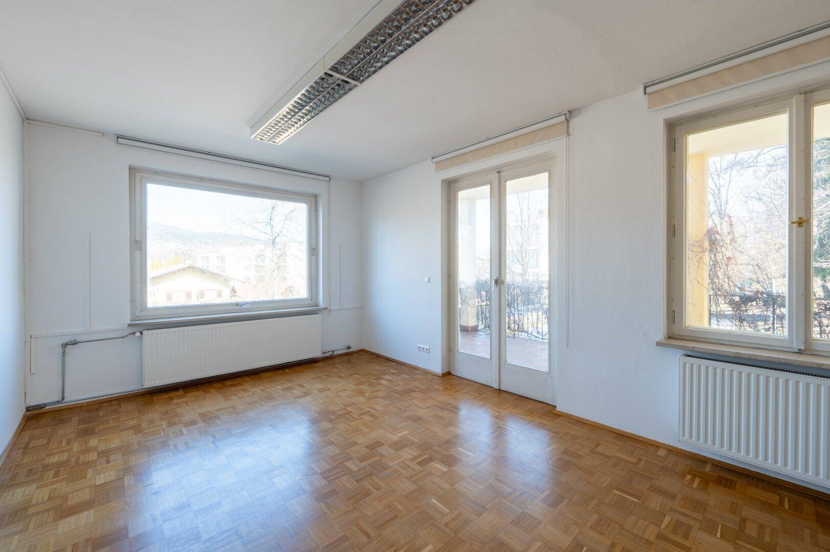Helles Zimmer mit Fischgrätparkett, großen Fenstern und Zugang zu einem Balkon.