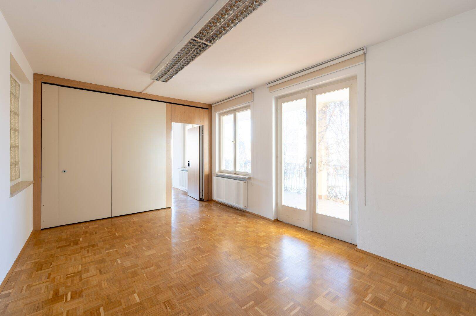 Heller Raum mit Parkettboden, Schiebetüren und großen Fenstern, die viel Tageslicht hereinlassen.