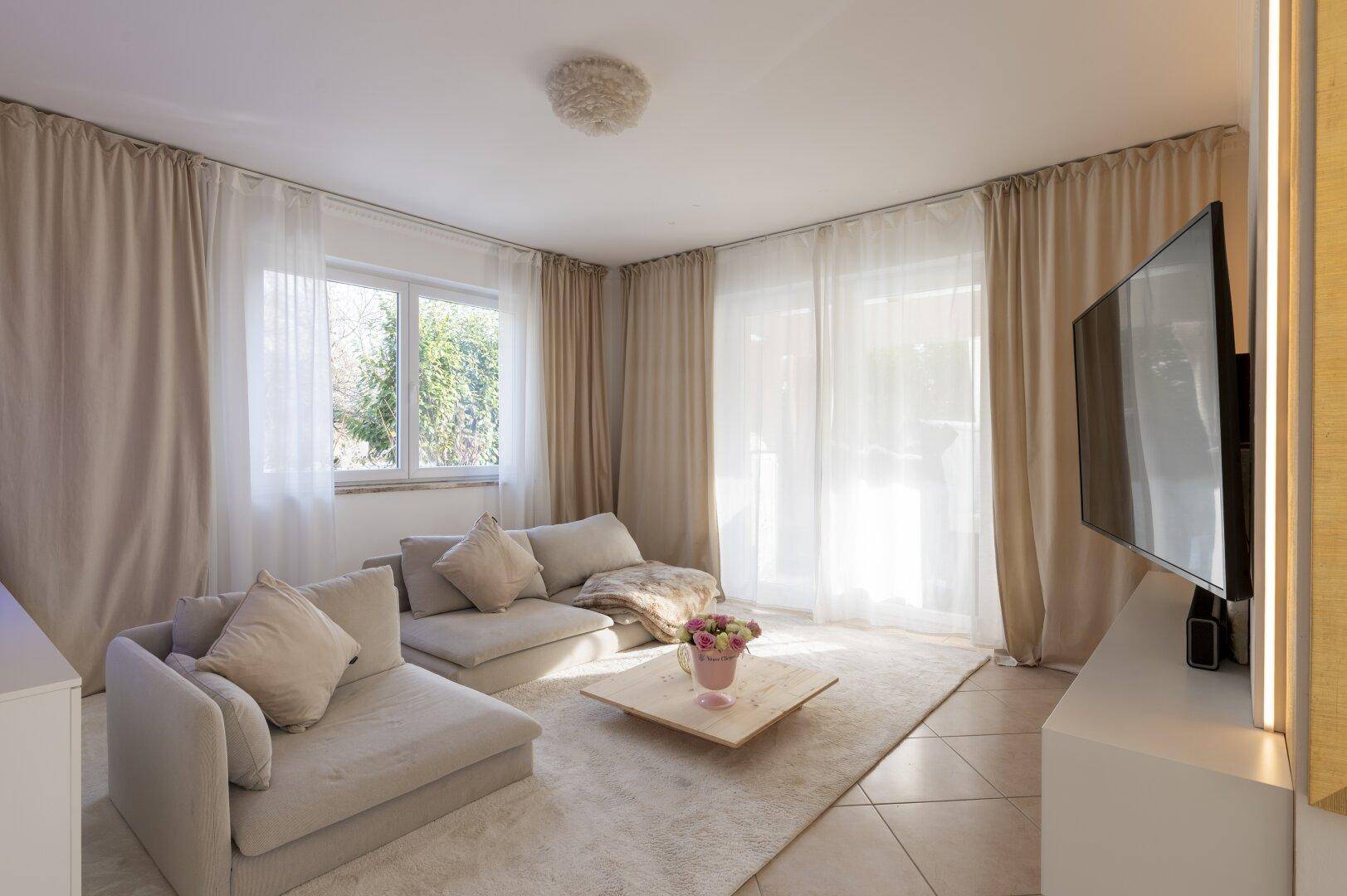 Helles Wohnzimmer mit bequemen Sofas, einem Fernseher und großen Fenstern.