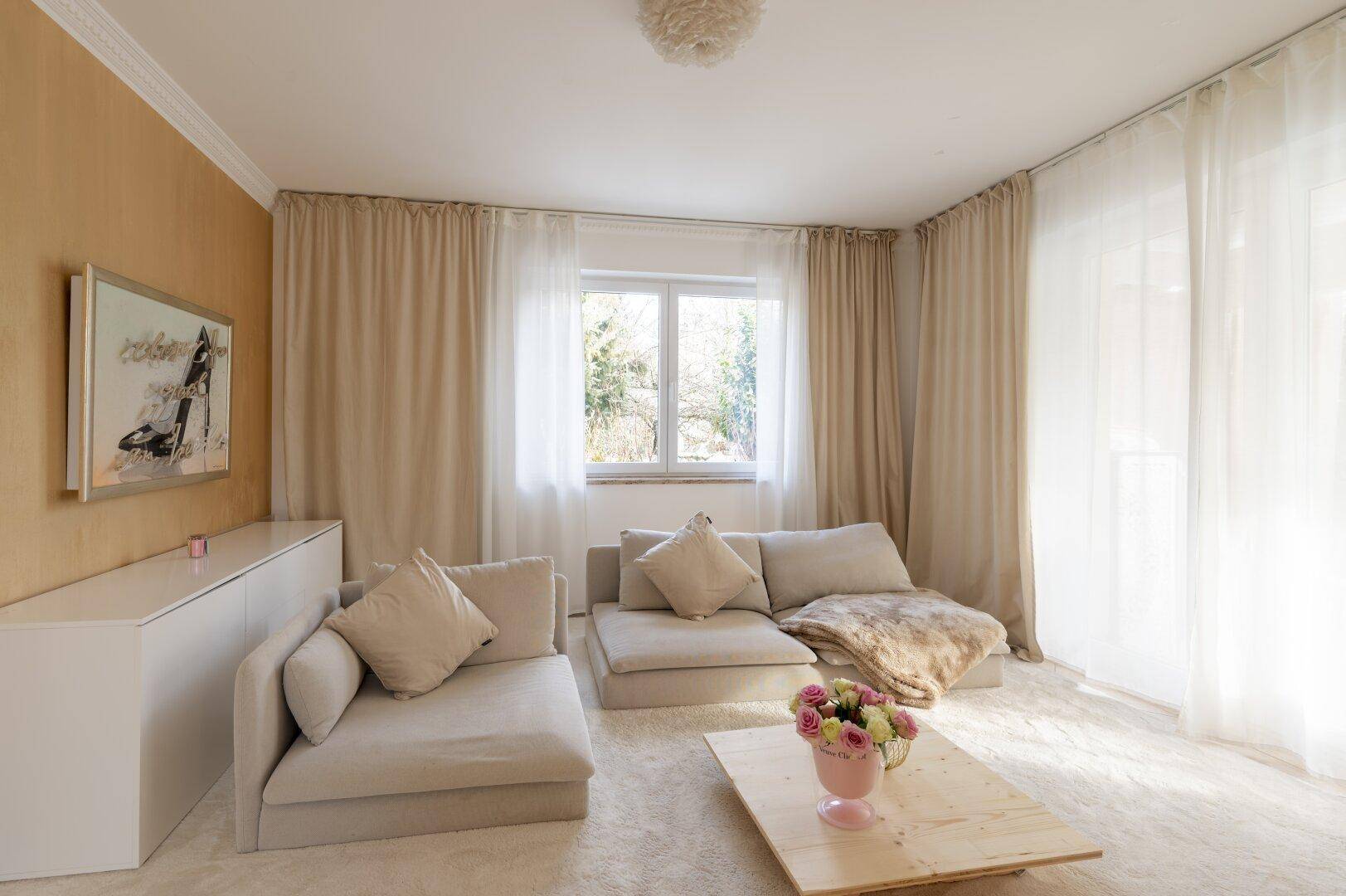 Gemütliches Wohnzimmer mit beigen Sofas, hellen Vorhängen und viel natürlichem Licht.