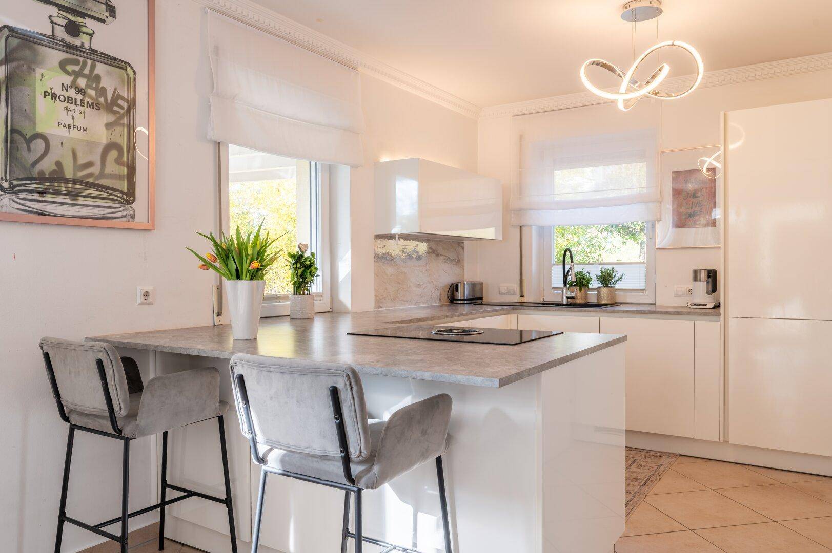 Moderne weiße Küche mit Kochinsel, Barhockern und viel Tageslicht durch die Fenster.