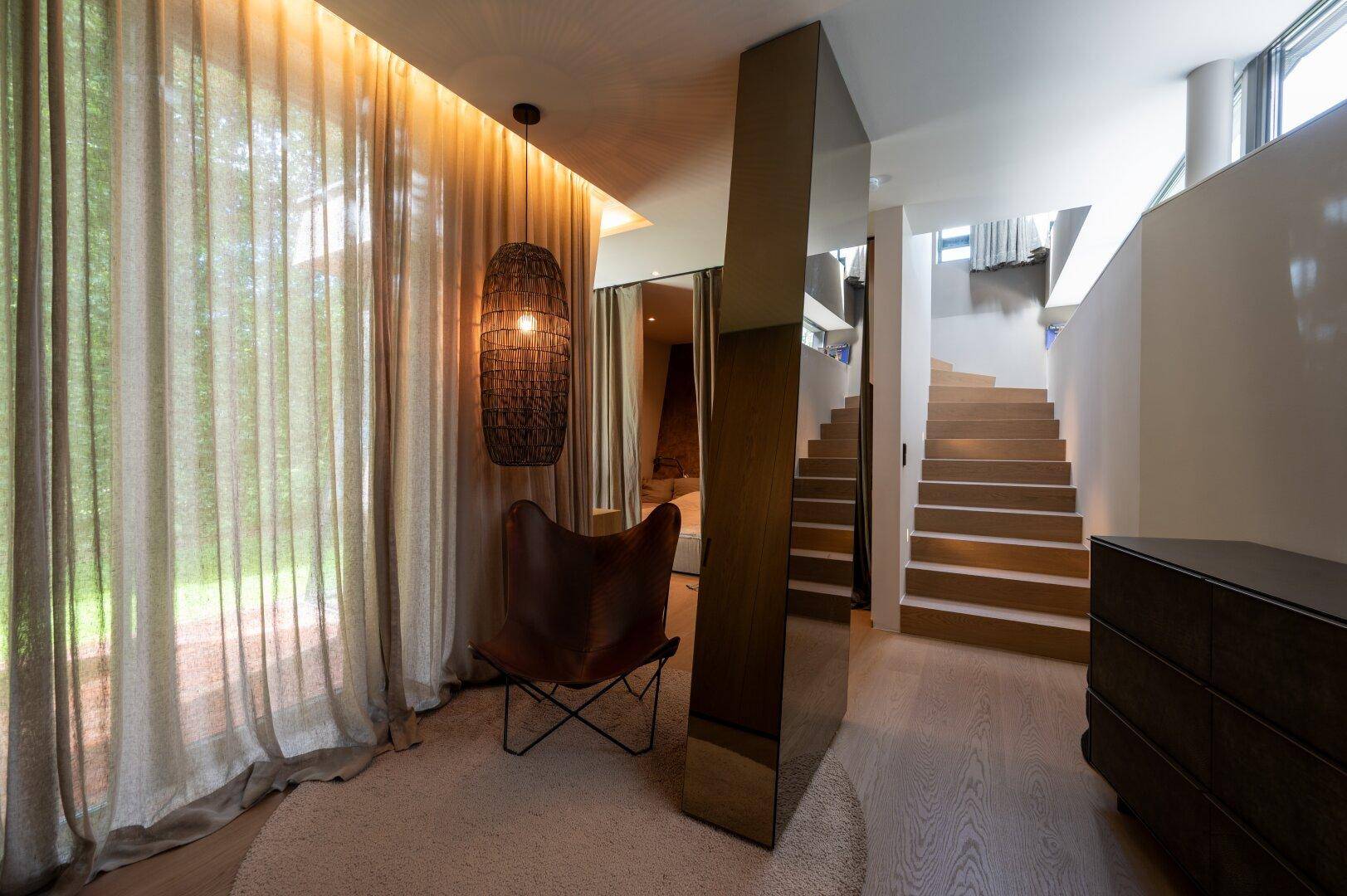 Heller Flurbereich mit eleganter Holztreppe, modernem Sessel und großen Fenstern mit Vorhängen.