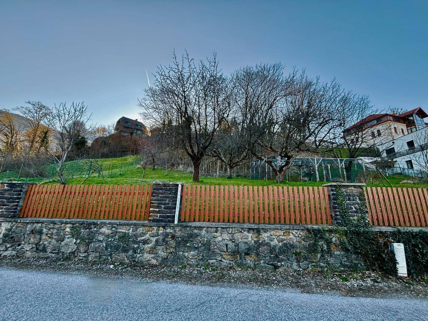 Holzzaun auf einer Natursteinmauer mit kahlen Bäumen und einem Haus im Hintergrund.