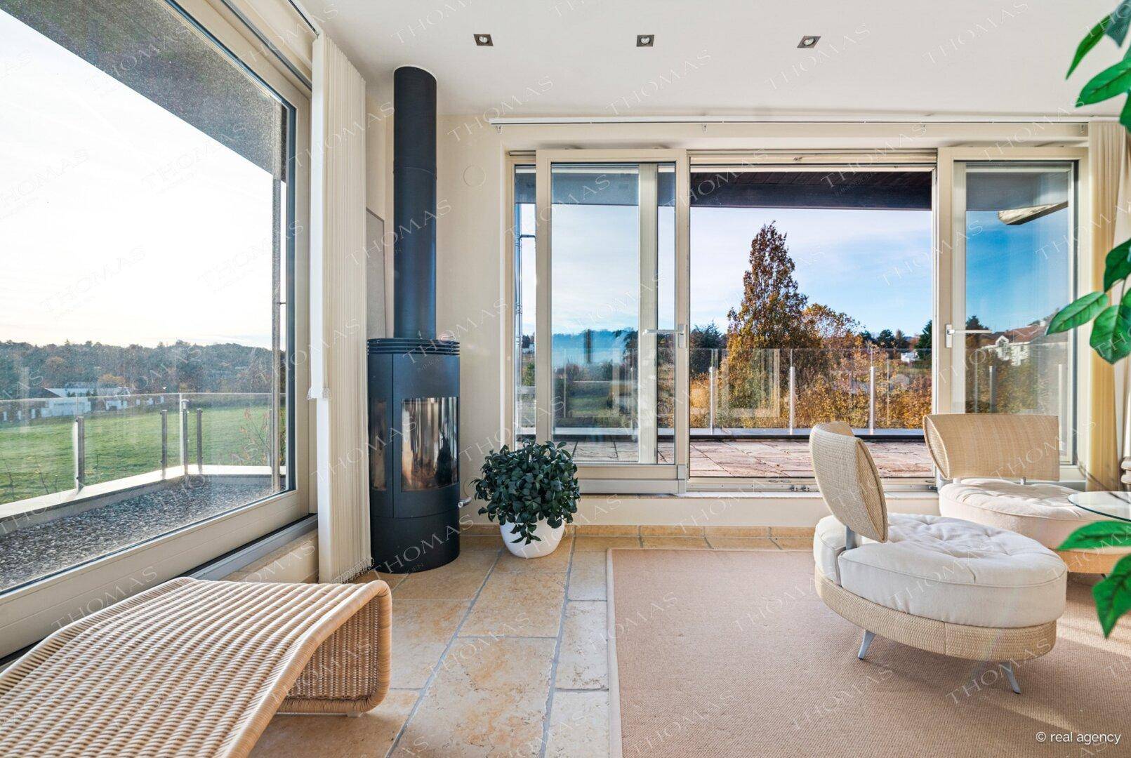 Helles Wohnzimmer mit modernem Kaminofen, Fliesenboden und Panoramablick ins Grüne.