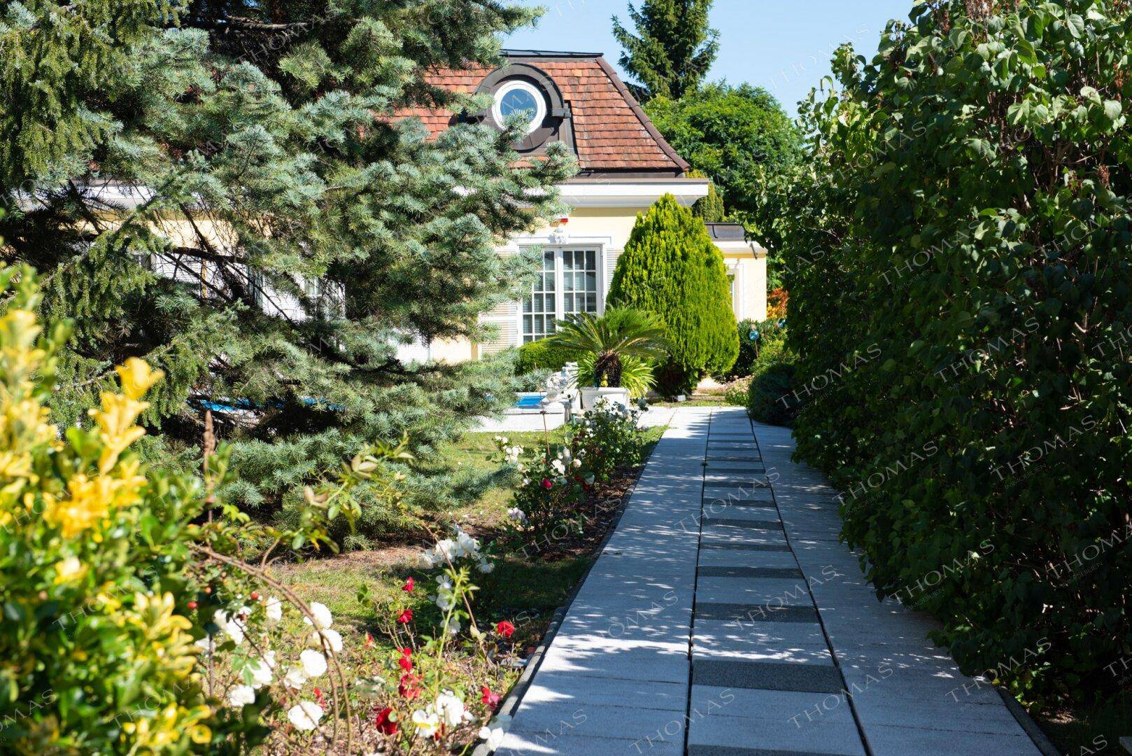 Gepflasterter Gartenweg führt durch üppiges Grün zum charmanten Haus mit Mansarddach.
