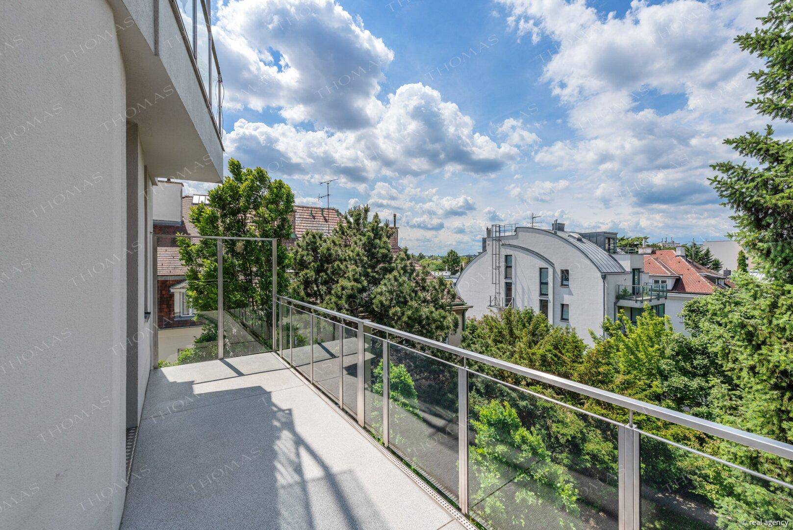 Geräumiger Balkon mit Glasgeländer und weitem Blick über grüne Bäume und die Umgebung.
