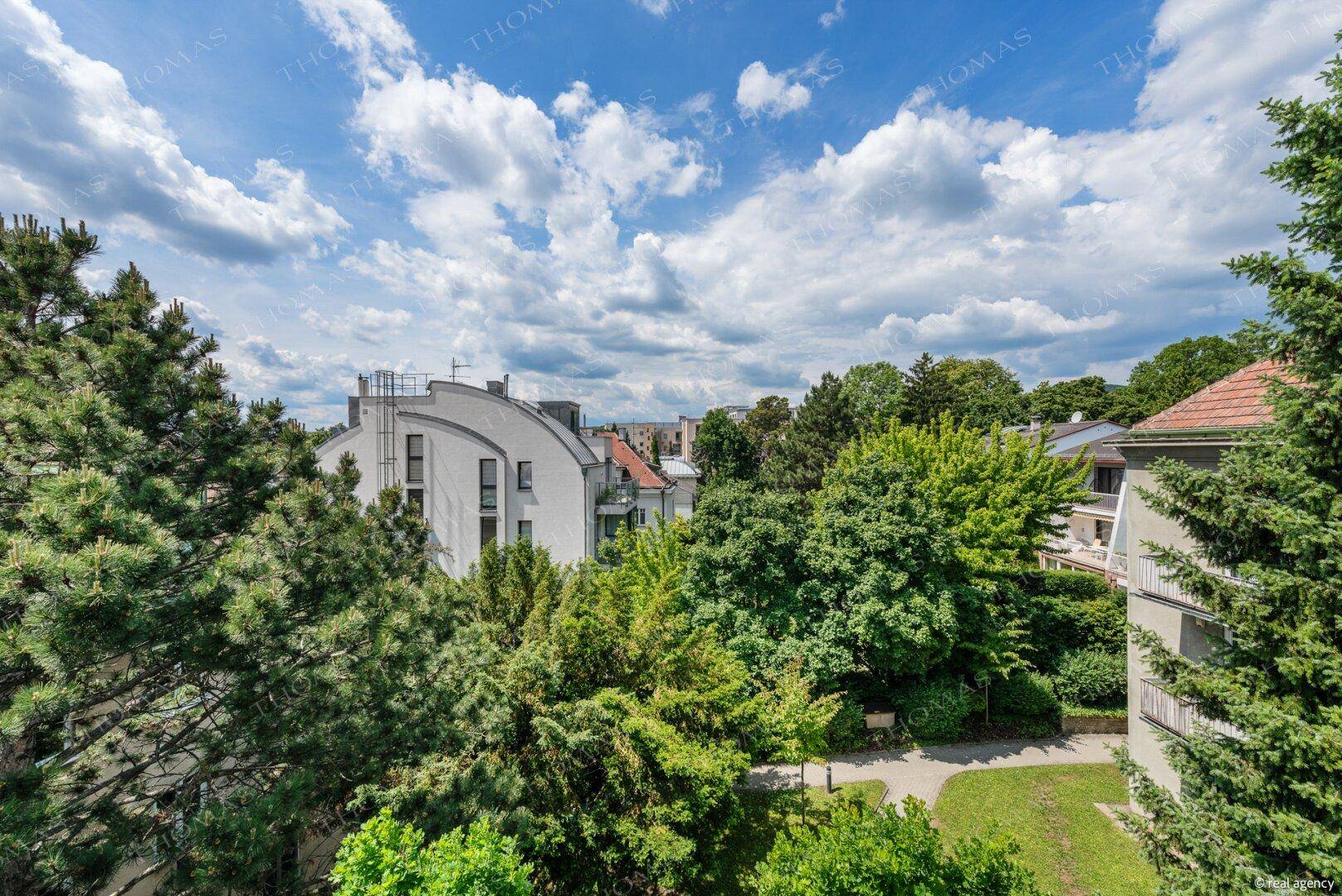 Weitläufiger Blick vom Balkon auf grüne Baumwipfel und die Dächer der umliegenden Gebäude.