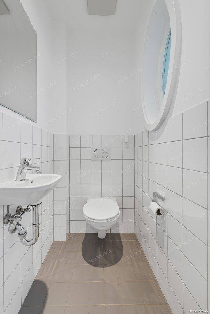 Kleines Gäste-WC mit weißen Fliesen, modernem WC und einem einzigartigen ovalen Fenster.