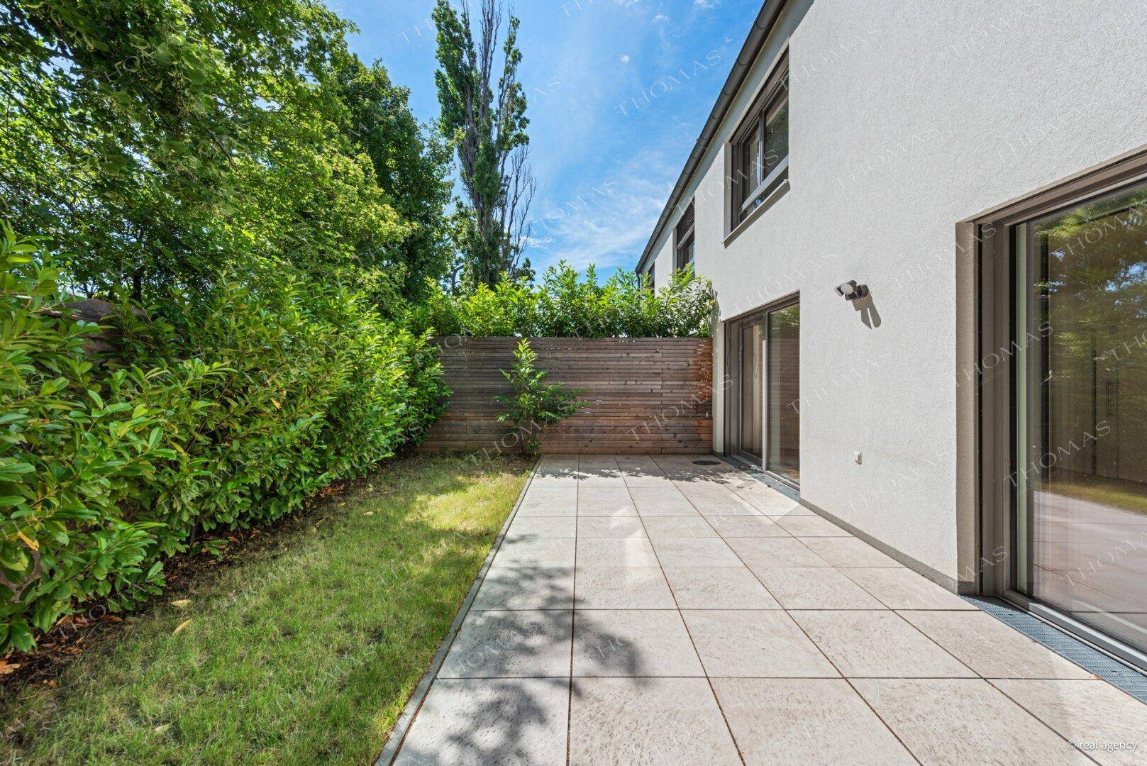 Sonniger Terrassenbereich mit hellen Fliesen, umgeben von einer grünen Hecke und Holzzaun.