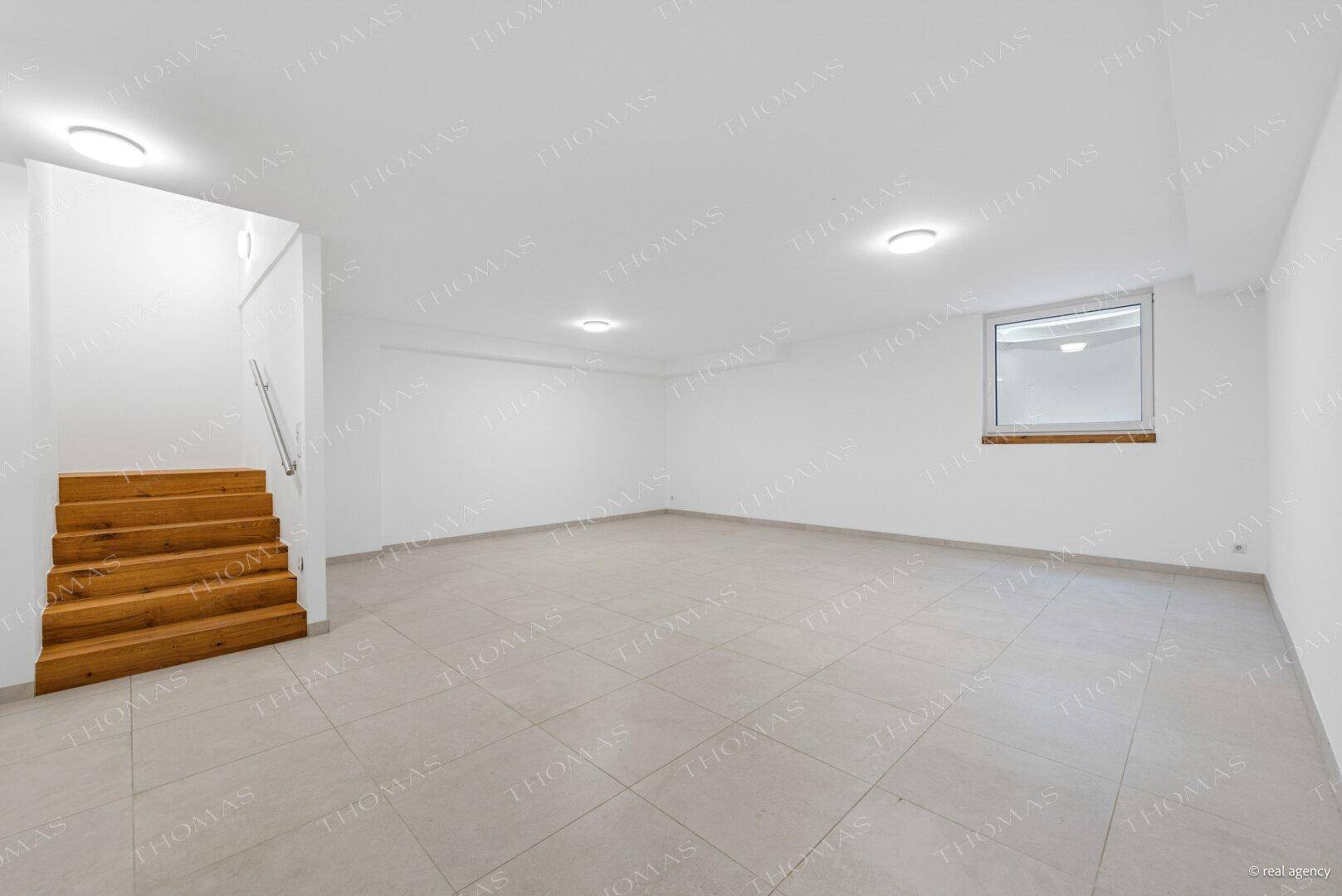 Geräumiger Kellerraum mit Fliesenboden, Holztreppe und einem Fenster, das Tageslicht hereinlässt.