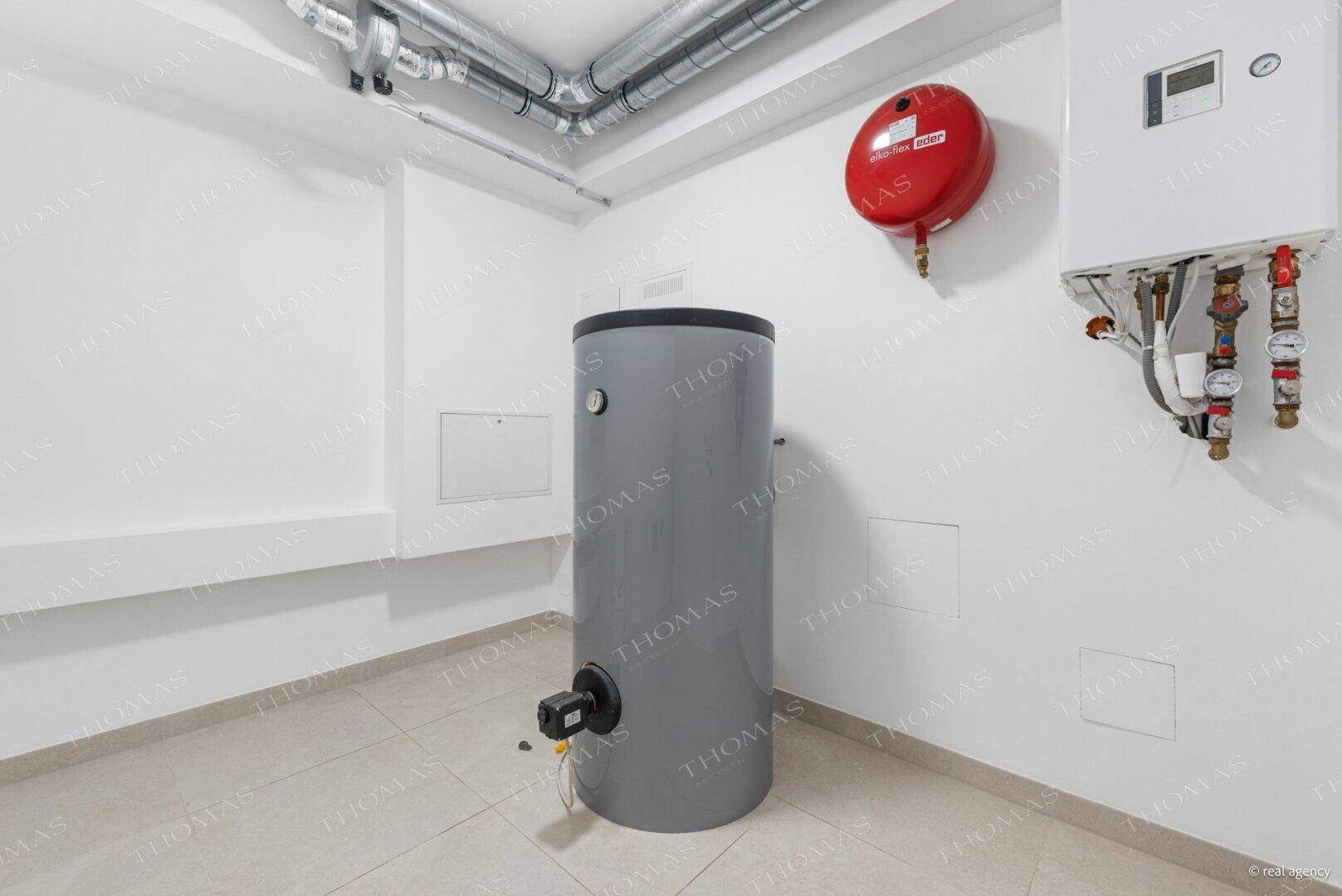 Technikraum mit Warmwasserspeicher, Heizungsanlage und sichtbaren Rohrleitungen an der Decke.