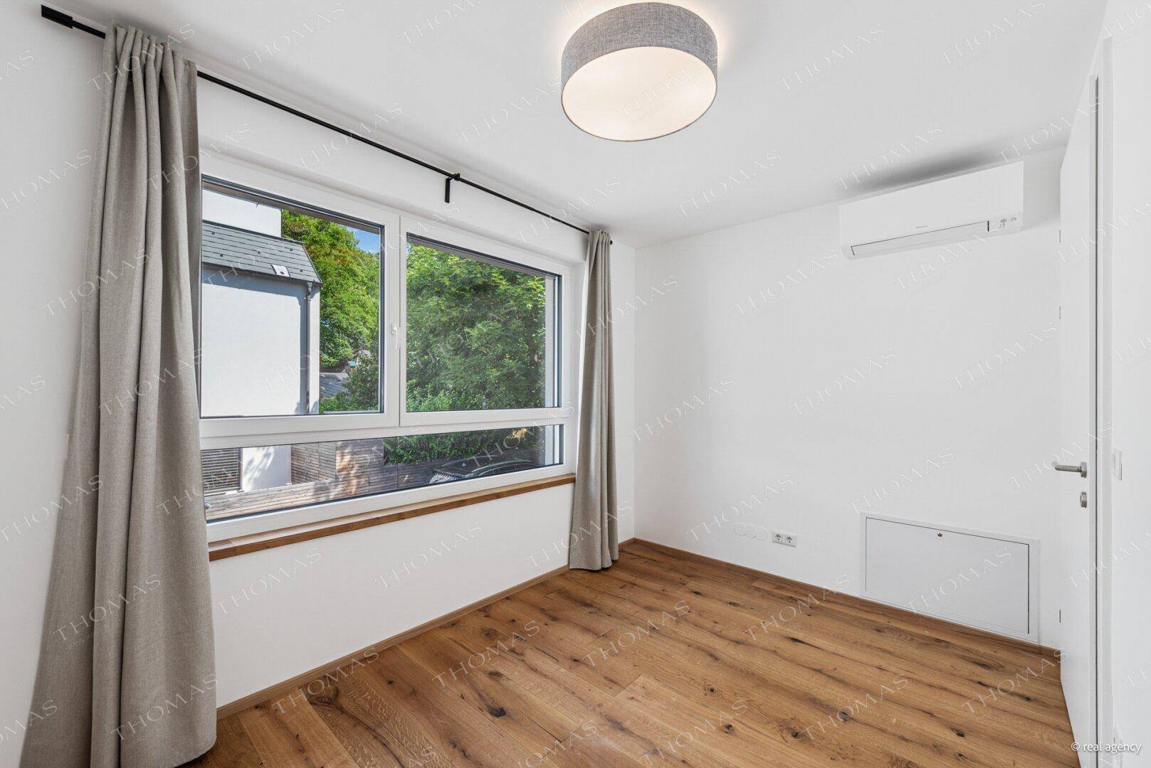 Helles Schlafzimmer mit Holzboden, Vorhängen und Klimaanlage, Blick ins Grüne.