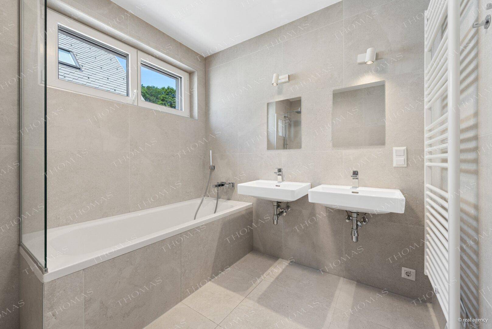 Modernes Badezimmer mit grauen Fliesen, Badewanne, Doppelwaschbecken und Handtuchheizkörper.