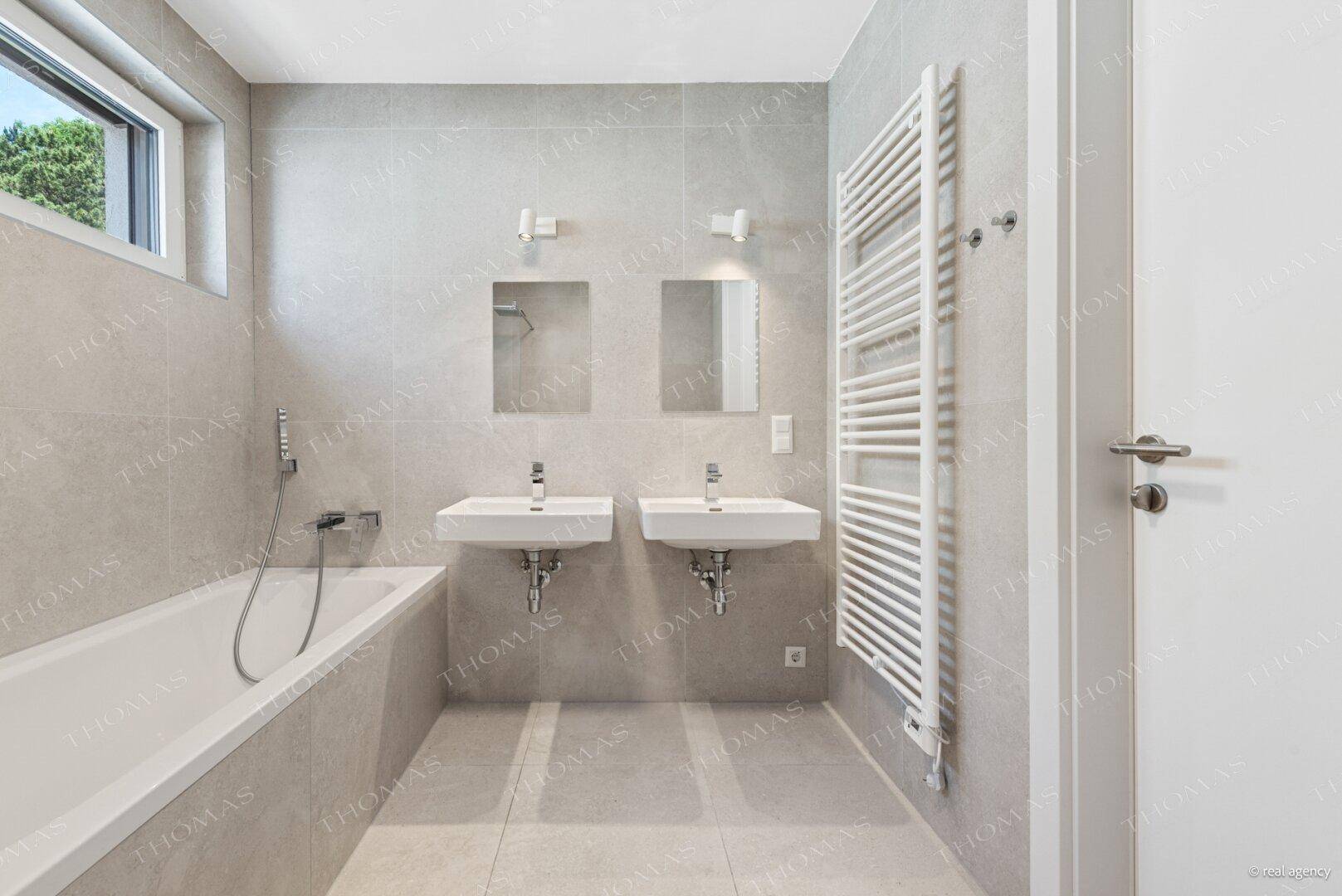Helles Badezimmer mit grauen Fliesen, Badewanne, Doppelwaschbecken und Handtuchheizkörper.
