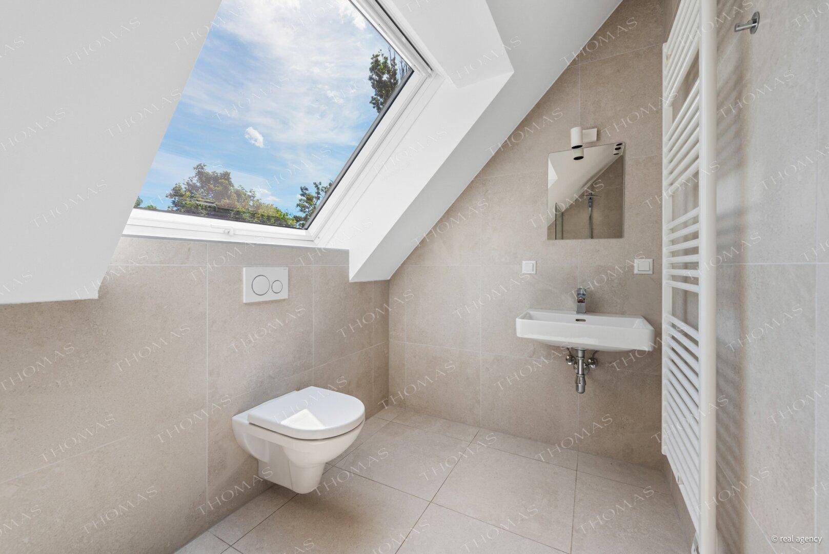 Modernes WC mit grauen Fliesen, wandhängender Toilette, Waschbecken und Dachfenster.