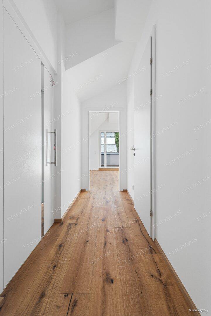 Heller Flur mit Holzboden und schräger Decke, der zu einem Zimmer mit Balkon führt.