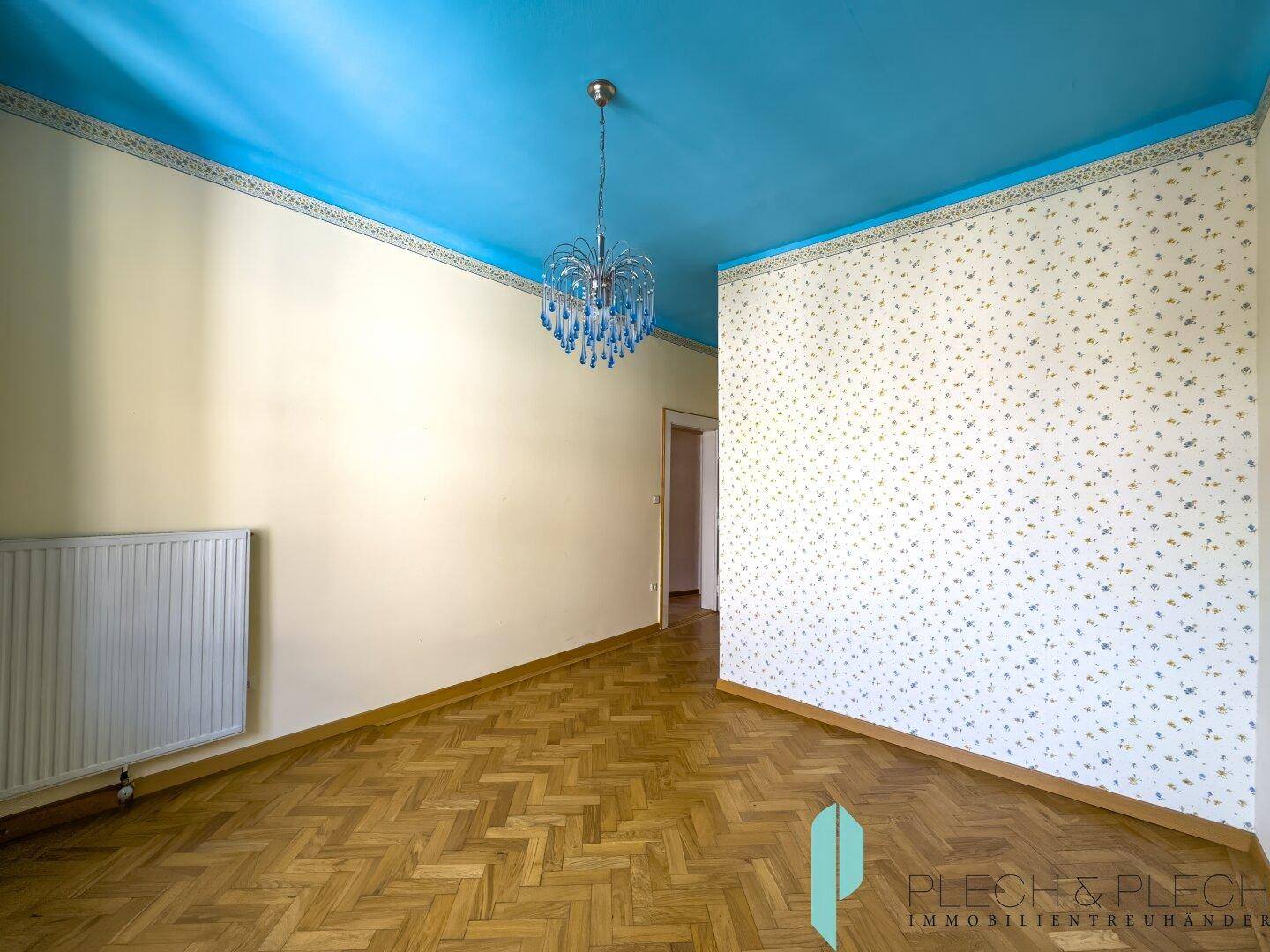 Helles Zimmer mit blauer Decke, Fischgrätparkett und gemusterter Tapete.