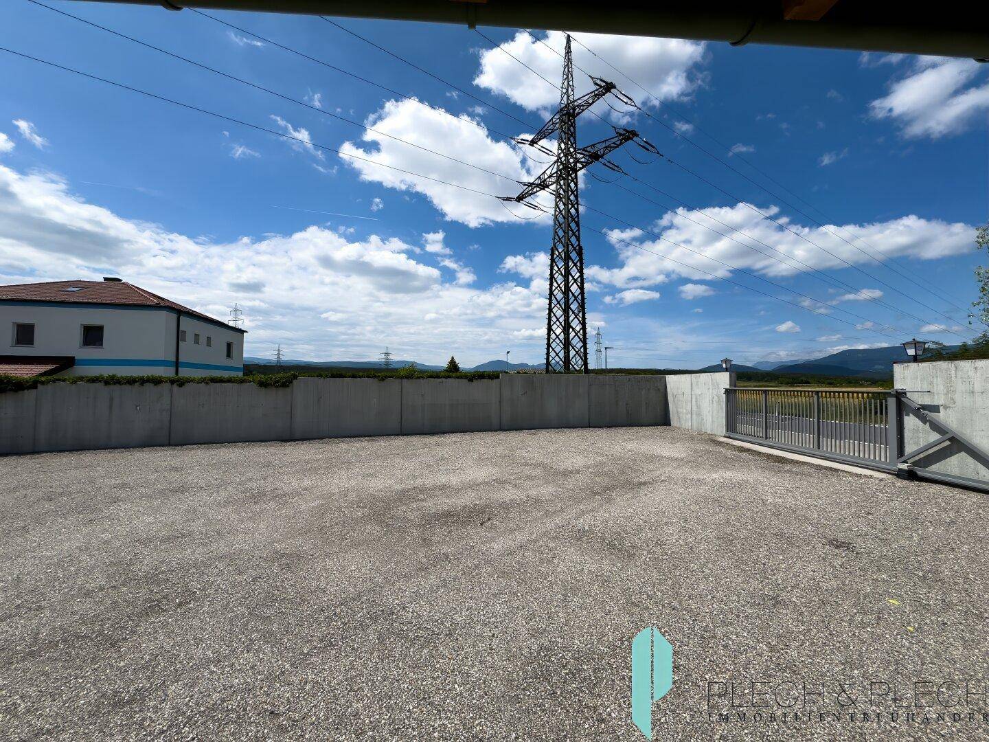 Großer Schotterbereich mit Betonmauer, Tor und Strommast vor einer Bergkulisse.