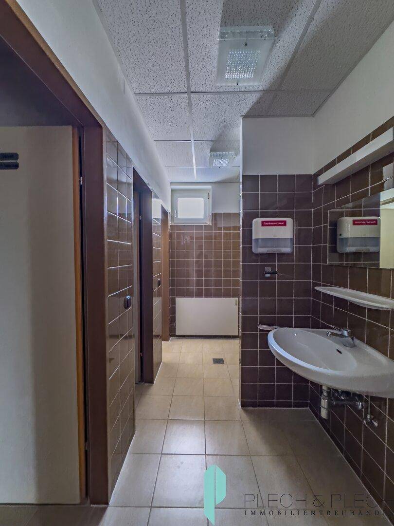 Flur im Sanitärbereich mit Waschbecken und braunen Fliesen, führt zu weiteren Toiletten.