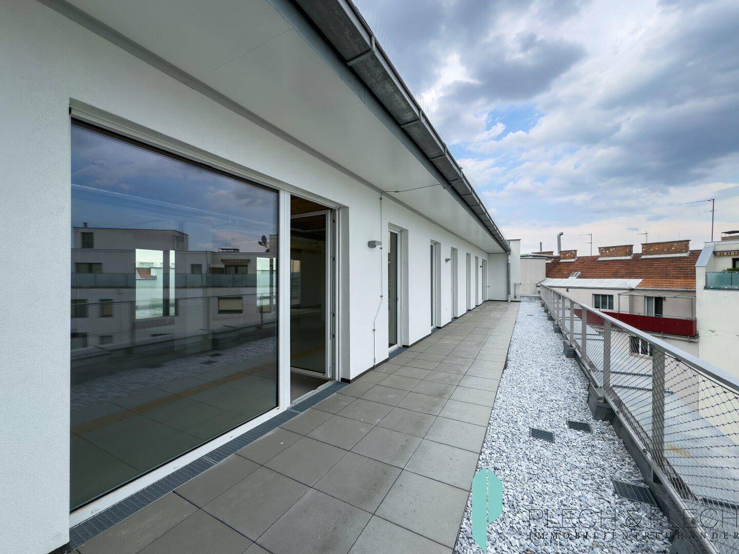 Lange Dachterrasse mit grauen Fliesen und Kiesstreifen, umgeben von modernen Gebäudefassaden unter bewölktem Himmel.