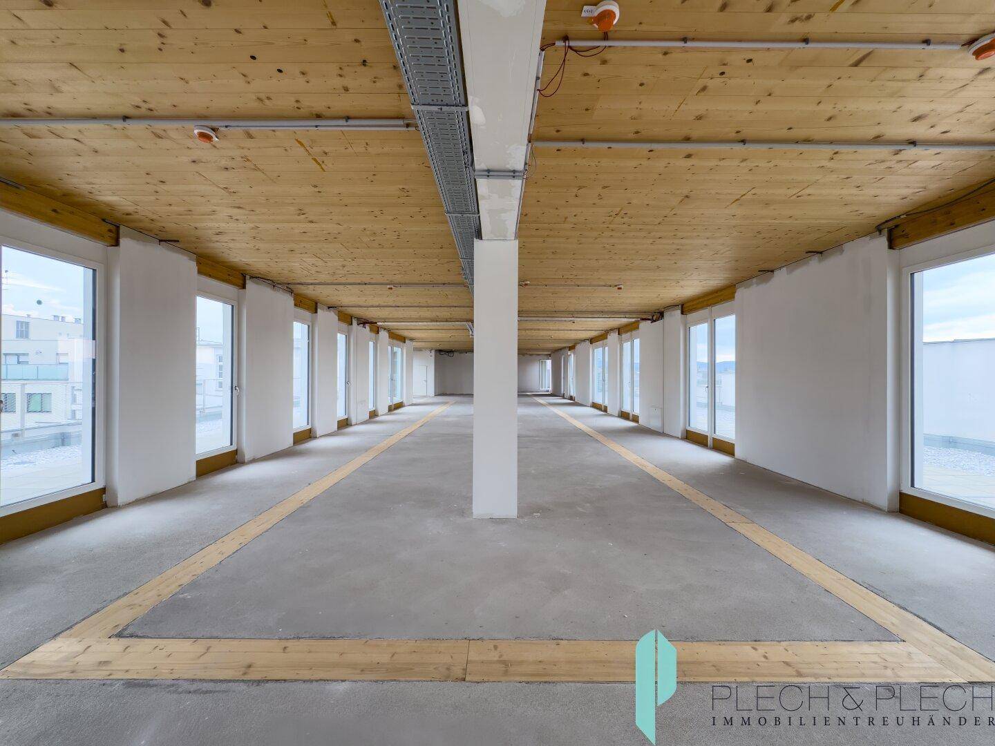 Weitläufiger Gewerberaum mit sichtbaren Holzbalken an der Decke und einer Reihe von Fenstern, die viel Tageslicht hereinlassen.
