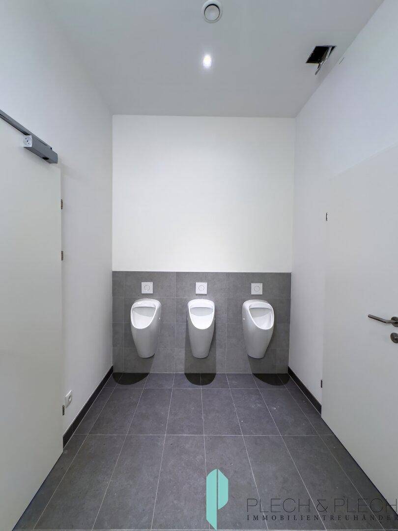 Moderner Herrenwaschraum mit drei Urinalen, grauen Fliesen und weiß gestrichenen Wänden.