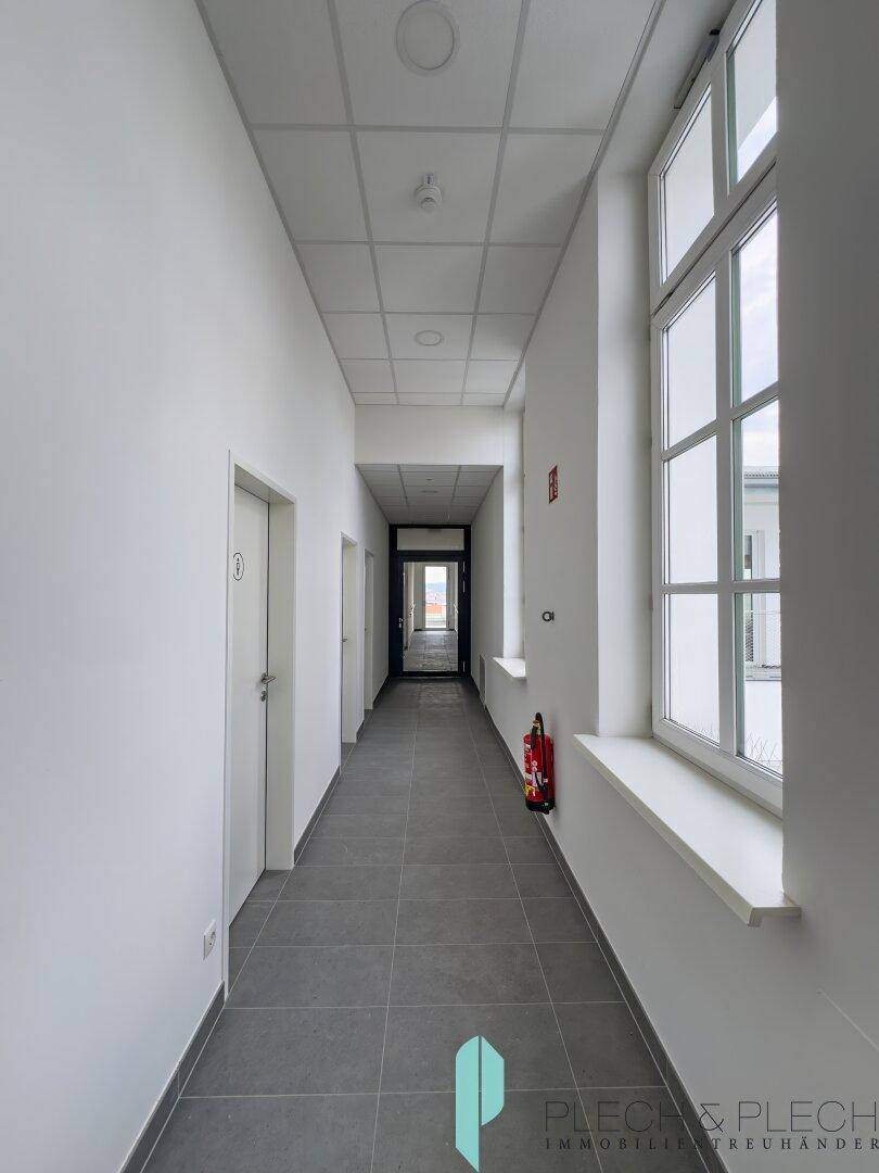 Langer, heller Flur mit grauen Bodenfliesen und großen Fenstern, die viel Tageslicht hereinlassen.