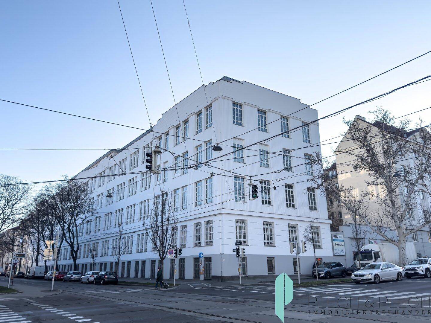 Großes, weißes Gebäude mit vielen Fenstern an einer städtischen Kreuzung unter blauem Himmel.