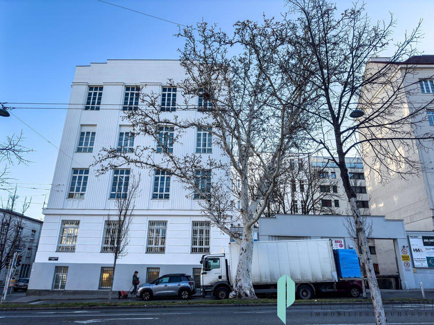 Weißes Gebäude mit vielen Fenstern und einem großen Baum davor, an einer städtischen Straße gelegen.